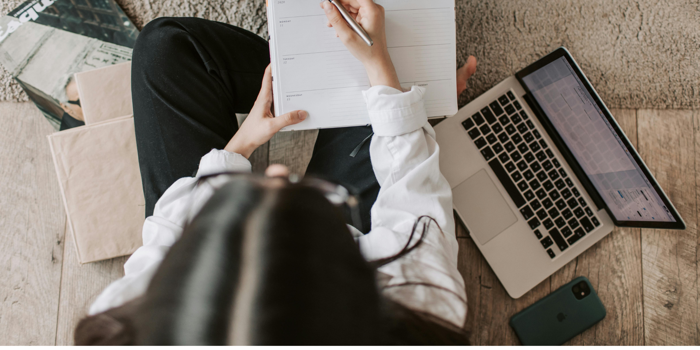 Woman sitting cross-legged on a couch writing in a schedule with Apple Mac-Book placed next to her