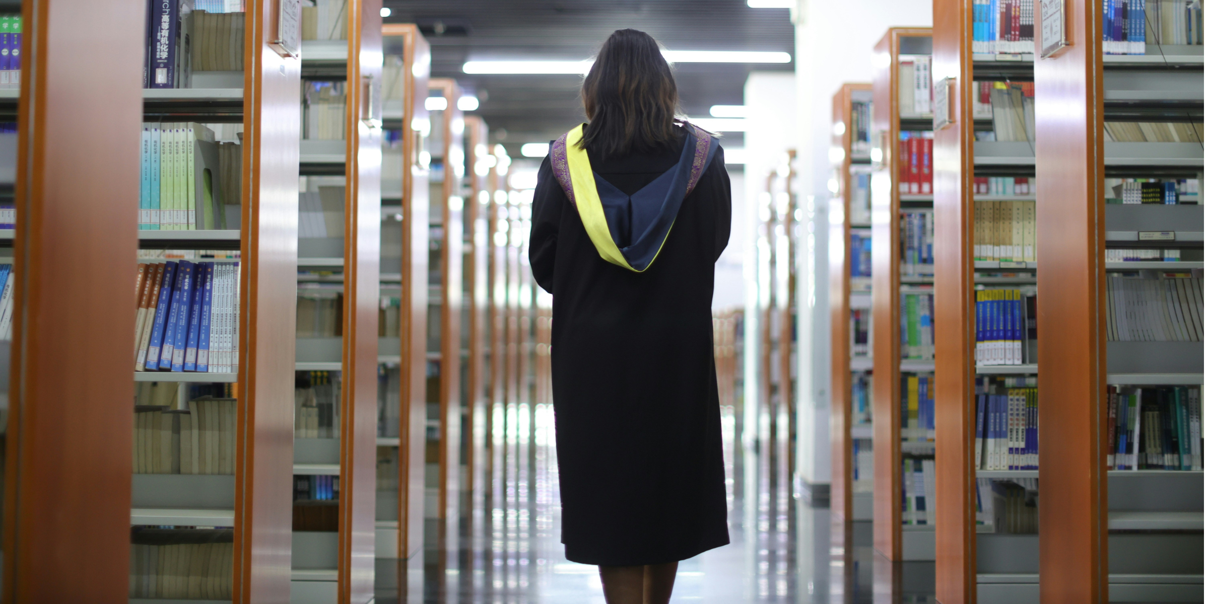 A woman in a graduation gown standing still in a library looking ahead, over-the-shoulder shot